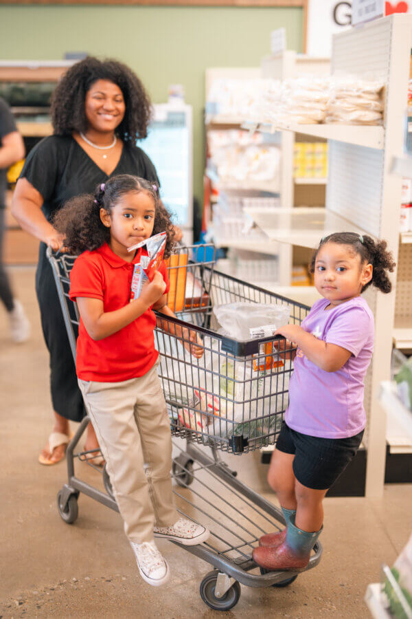 Turnip Truck & 'The Store' Are Rounding Up for Neighbors in Need - 2 A woman and two young girls stand with a full shopping cart in the store aisle. One girl is holding snacks, while the other stands inside the cart, looking like she just hopped off the turnip truck for a fun grocery adventure.