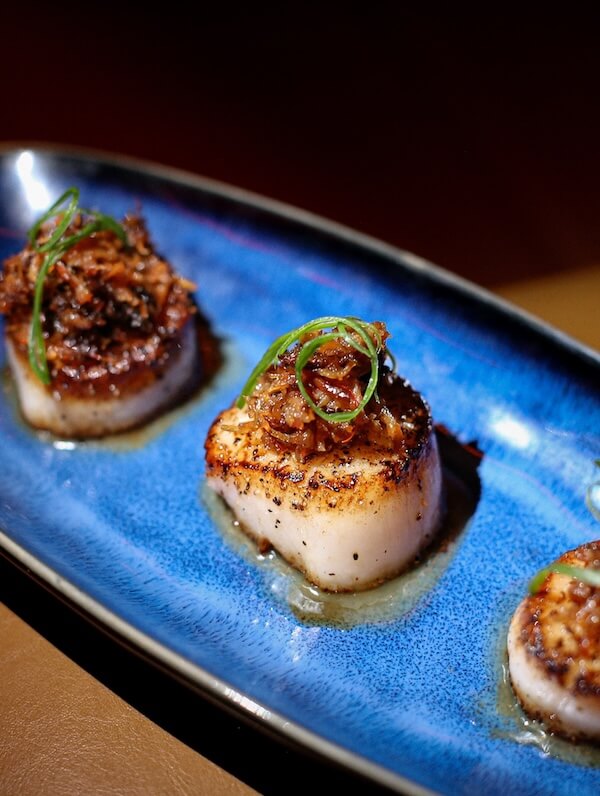 Sushi in Nashville is a THING. Here's Your Guide. - 3 Three seared scallops topped with caramelized onions and green garnish are arranged on a blue oval plate, offering a Nashville twist on seafood elegance reminiscent of refined sushi presentations.