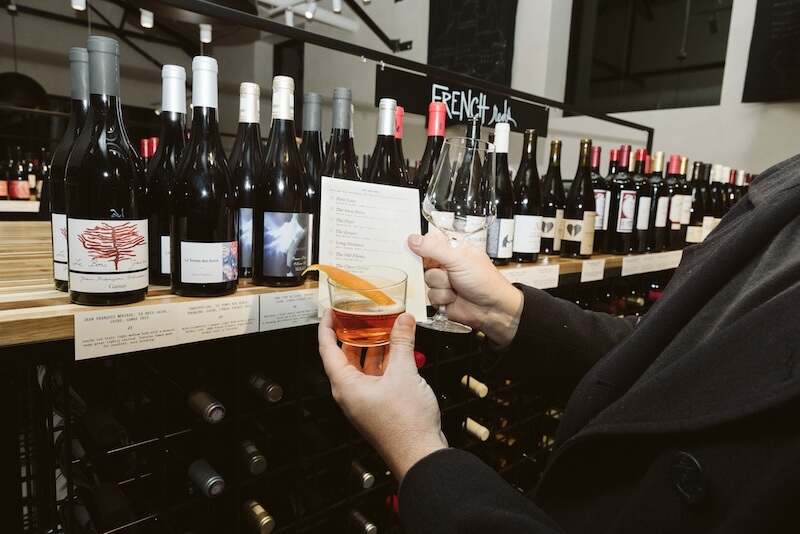 9 New Nashville Retail Spots to Explore - 4 Person holding a wine glass and a tasting menu in front of shelves stocked with various bottles of wine at a new retail spot in Nashville.