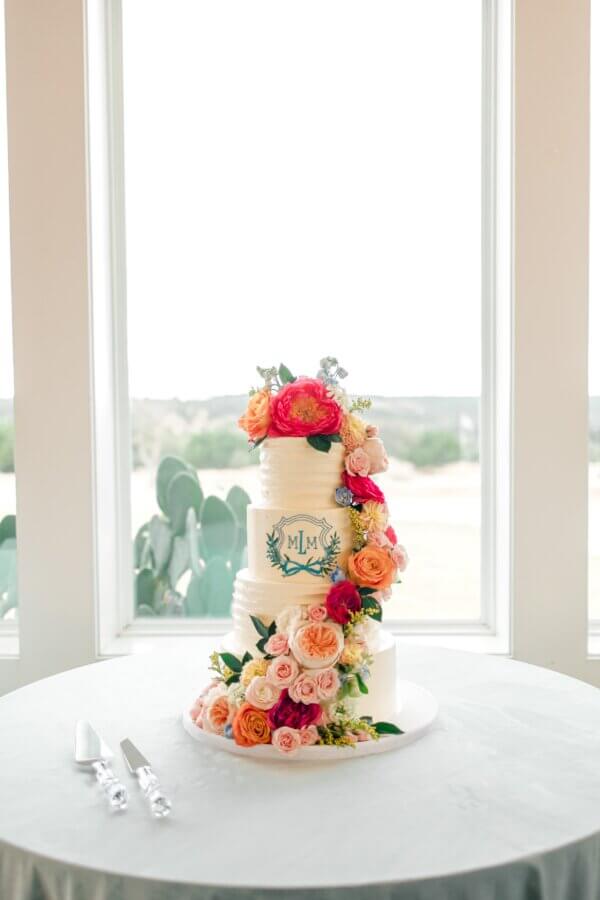 Rustic Meets Romance in This Texas Hill Country Wedding - 15 A two-tier white wedding cake decorated with colorful fresh flowers and a monogram sits on a round table in front of large windows—perfect for a romantic Fredericksburg, Texas wedding. Cake-cutting utensils are placed on the table.