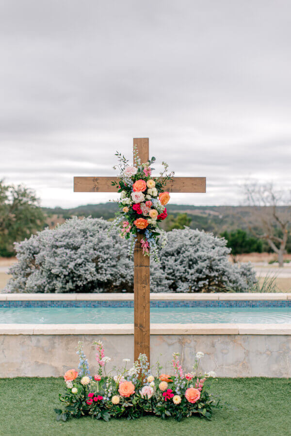 Rustic Meets Romance in This Texas Hill Country Wedding - 12 A wooden cross decorated with colorful flowers stands in front of a small pool and landscaped shrubbery under a cloudy sky, capturing the charm of a Hill Country Wedding in Fredericksburg, Texas.