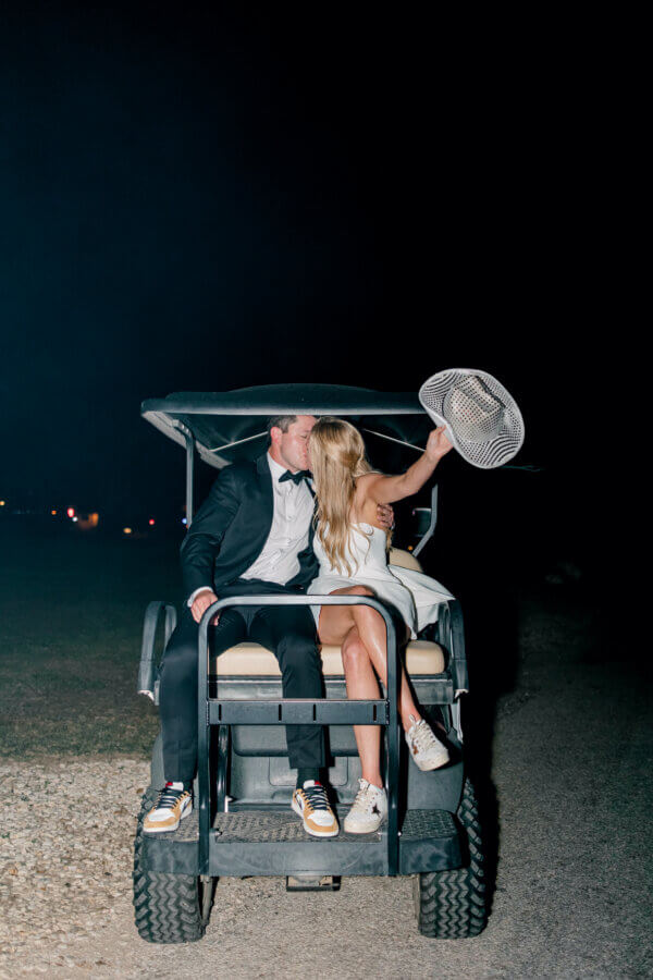 Rustic Meets Romance in This Texas Hill Country Wedding - 19 A couple dressed in formal attire sits on the back of a golf cart at night, kissing. The woman holds a wide-brimmed hat in the air, capturing the romance of a Hill Country wedding under the Texas stars.