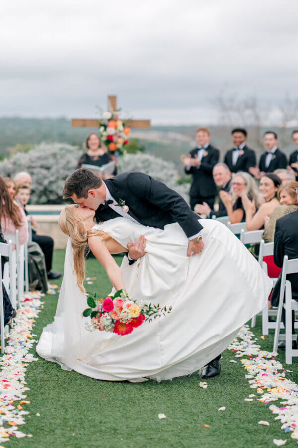 Rustic Meets Romance in This Texas Hill Country Wedding - 2 A bride and groom kiss as the groom dips the bride at an outdoor Hill Country wedding, surrounded by guests seated along a petal-strewn aisle.