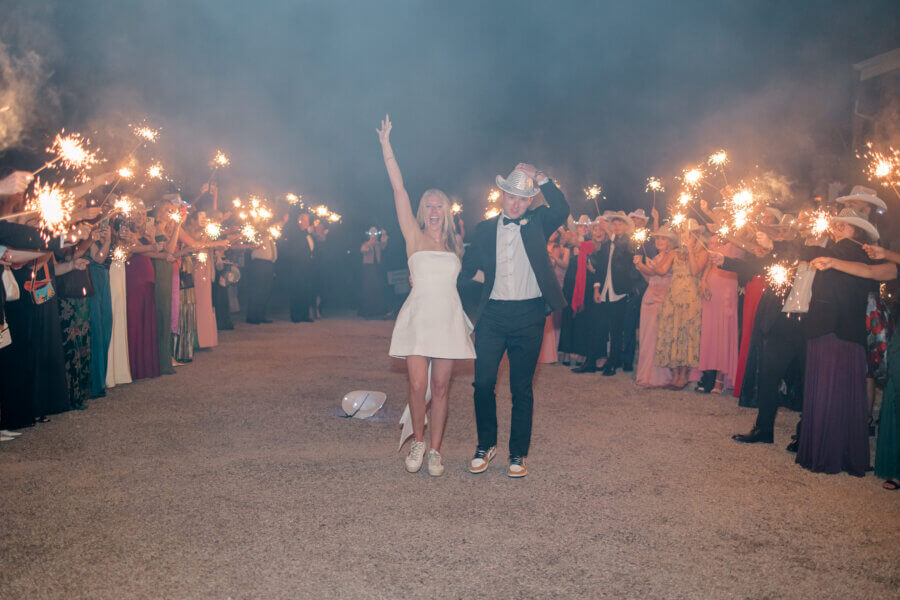 Rustic Meets Romance in This Texas Hill Country Wedding - 18 A couple in formal attire walks down a path lined with guests holding sparklers at night during a joyful Hill Country wedding; the woman raises her arm as they celebrate under the Texas sky.