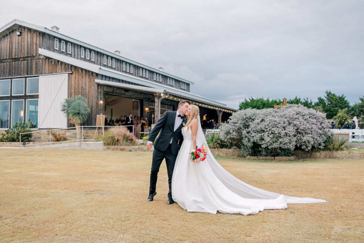 A bride and groom kiss outdoors in front of a rustic barn venue during their romantic wedding; the bride holds a bouquet and wears a long white gown with a train.