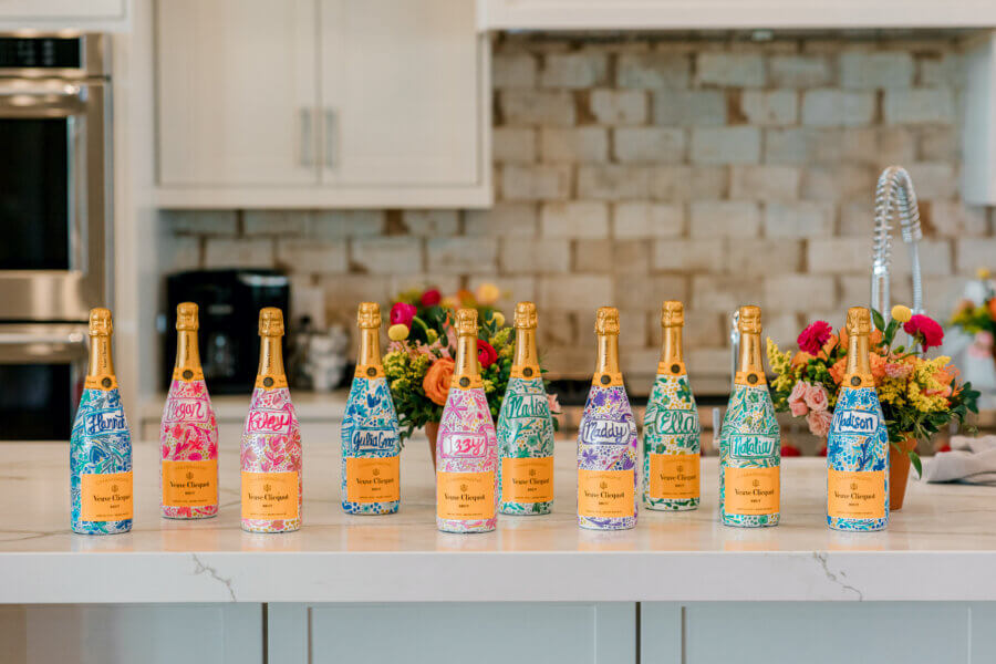 Rustic Meets Romance in This Texas Hill Country Wedding - 9 Ten champagne bottles with colorful, personalized labels are lined up on a white kitchen island, with elegant flower arrangements in the background.