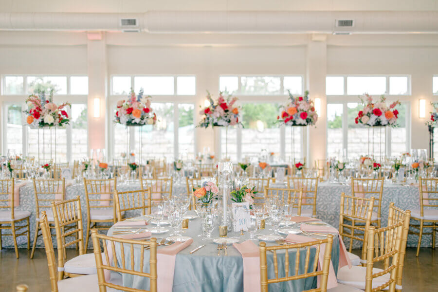 Rustic Meets Romance in This Texas Hill Country Wedding - 14 Round tables with gold chairs, set for a Hill Country wedding, feature pink and white floral centerpieces in a bright room with large windows.