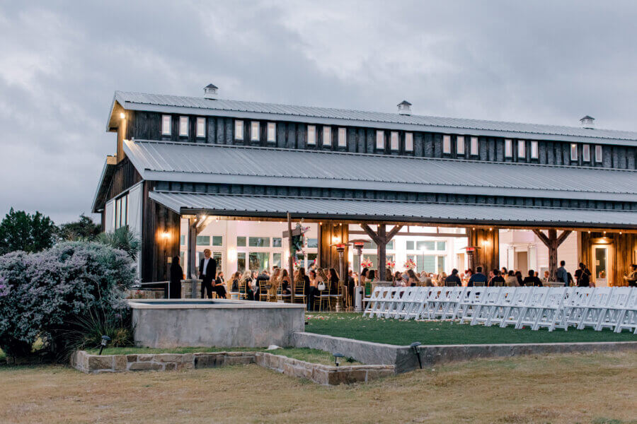 Rustic Meets Romance in This Texas Hill Country Wedding - 4 A large barn-style building in Fredericksburg, Texas hosts an event with people seated inside and empty white chairs arranged outside under a covered patio.