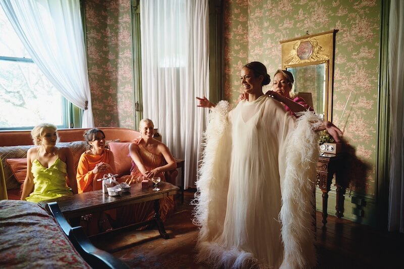 A Dreamy Disco Wedding at The Columns NOLA - 7 A woman in a white feathered gown stands with arms outstretched, exuding disco wedding glamour, while three women sit on a sofa and another stands behind her in a vintage-styled room.