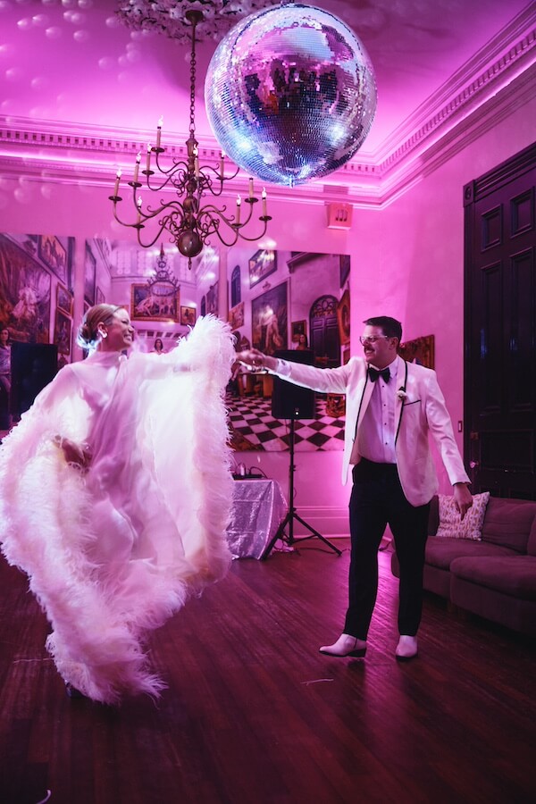 A Dreamy Disco Wedding at The Columns NOLA - 12 A couple in formal attire dances under a disco ball at their disco wedding, surrounded by pink lighting and elegant chandeliers.