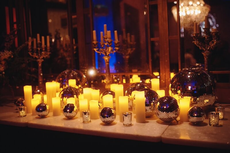 A Dreamy Disco Wedding at The Columns NOLA - 4 A marble surface decorated for a disco wedding, featuring lit pillar candles, silver disco balls, glass votives, and brass candelabras, with a mirror reflecting the warm candlelight.