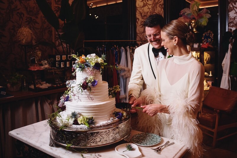 A Dreamy Disco Wedding at The Columns NOLA - 13 A couple in formal attire smiles as they cut a tiered wedding cake decorated with colorful flowers at a lively indoor disco wedding reception.
