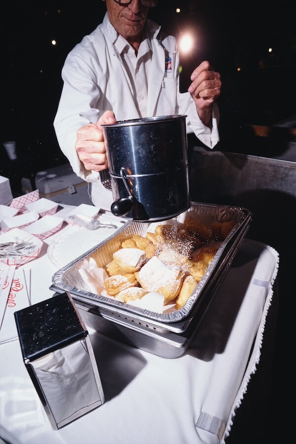 A Dreamy Disco Wedding at The Columns NOLA - 15 A person in a white coat sprinkles powdered sugar from a metal sifter onto pastries, preparing sweet treats for a disco wedding on a table covered with a white tablecloth.