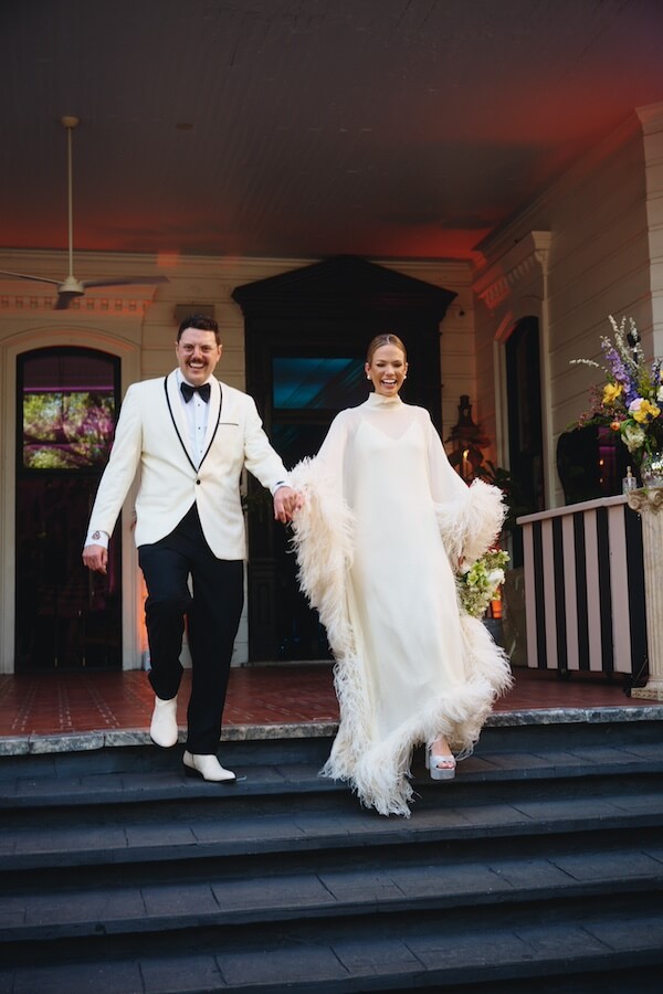 A Dreamy Disco Wedding at The Columns NOLA - 2 A couple in formal wedding attire walks down outdoor steps, smiling at their disco wedding; the bride wears a white dress with feathered sleeves, and the groom sports a white tuxedo jacket with black pants.
