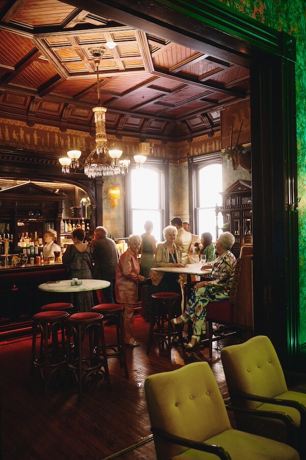 A Dreamy Disco Wedding at The Columns NOLA - 6 A group of elderly people sits and stands around a bar and high tables in a vintage-style room with ornate woodwork and a chandelier, celebrating what looks like a retro disco wedding.