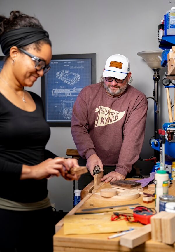 Kentucky's Kara Hill Turns Stem Cuttings Into Living Art - 8 Two people work on a woodworking project in a workshop, using various tools and materials on a cluttered workbench.