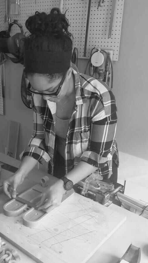 Kentucky's Kara Hill Turns Stem Cuttings Into Living Art - 7 A woman wearing glasses and a plaid shirt works on a woodworking project at a bench, using clamps and wooden pieces—tools for plant propagation hang on a pegboard in the background.