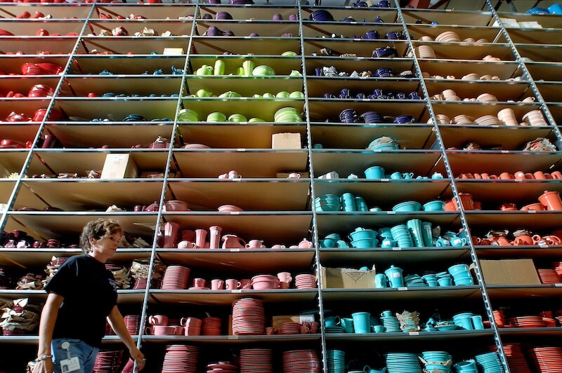 Where the "Good China" Lives: Inside Replacements, Ltd. - 5 A person walks past tall shelves filled with neatly organized, colorful ceramic dishware and Good China in various shades at Replacements Ltd.