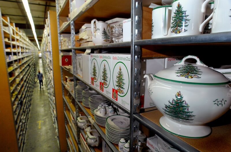 Where the "Good China" Lives: Inside Replacements, Ltd. - 4 Shelves stocked with Christmas-themed dishware, including Replacements Ltd Good China plates and teapots, line an aisle in a warehouse; a person walks in the distance.