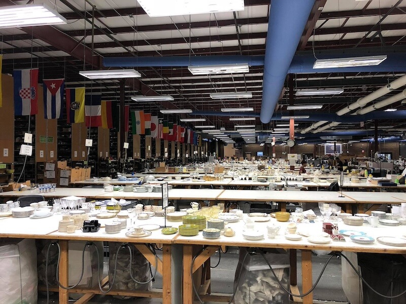 Where the "Good China" Lives: Inside Replacements, Ltd. - 2 Large warehouse with long tables covered in various dishes and kitchenware, including good china and unique finds from Replacements Ltd, surrounded by flags from different countries hanging from the ceiling.