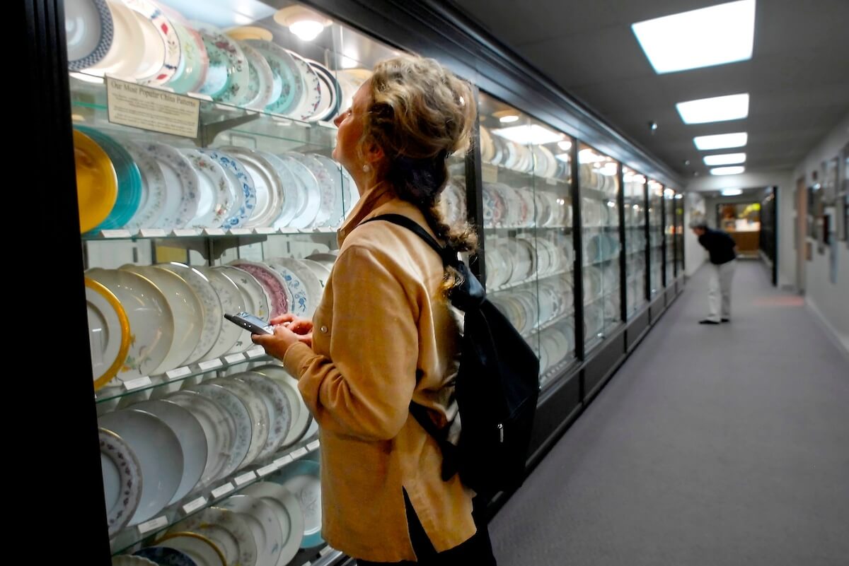 A woman with a backpack examines a display case filled with fine china plates in a well-lit museum or gallery hallway.