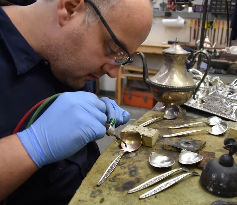 Where the "Good China" Lives: Inside Replacements, Ltd. - 6 A person wearing gloves and safety glasses uses a tool to repair a silver spoon at a workbench with various silverware, fine china, and tools—much like the restoration experts at Replacements Ltd.