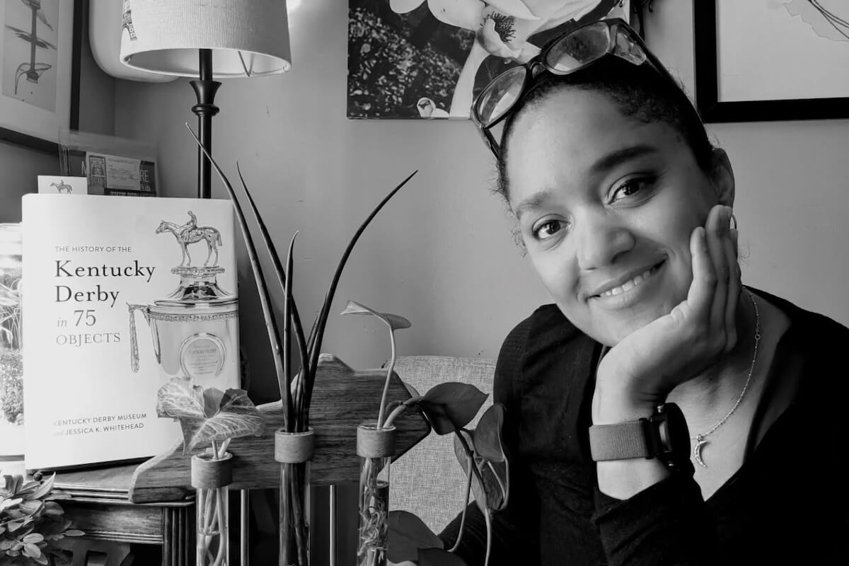 A woman smiles at the camera with her hand on her chin. A book titled "The History of the Kentucky Derby in 75 Objects" and some plants, hinting at plant propagation, are on the table beside her.