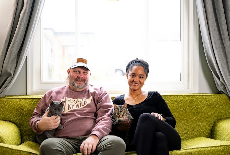 Kentucky's Kara Hill Turns Stem Cuttings Into Living Art - 6 Two adults sit on a green couch in front of a window, each holding a cat. The man wears a cap and sweatshirt, and the woman is in a black top, both smiling at the camera.