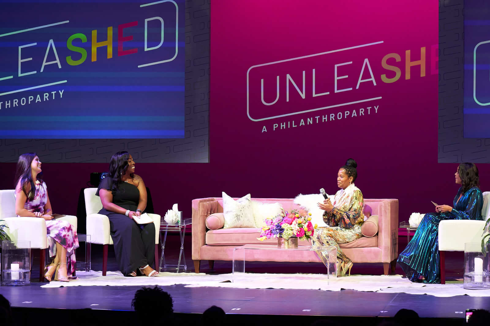 Four women sit on stage in conversation at the UNLEASHED philanthropy event, hosted by Red Mountain Theatre, with a pink backdrop and floral arrangements adorning the table.