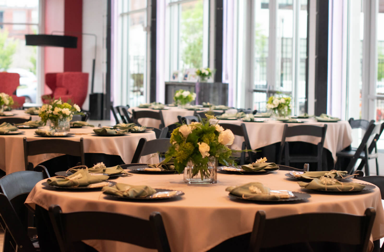 Round tables with white tablecloths, black chairs, and green floral centerpieces set for an event in the bright Red Mountain Theatre room with large windows.