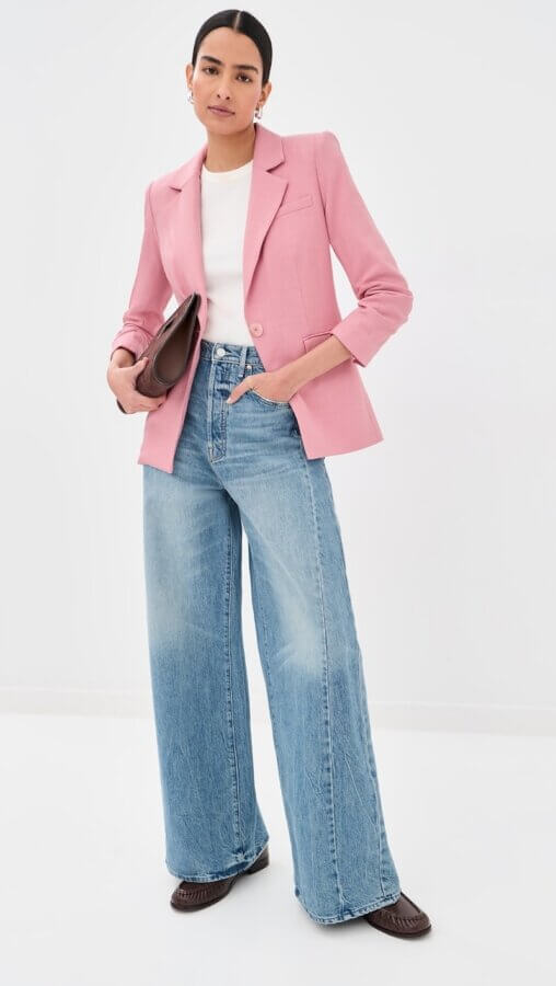 6 Spring 2026 Fashion Trends + How to Wear Them - 4 A person wearing a pink blazer, white top, wide-leg blue jeans, and brown shoes holds a brown clutch bag, standing against a plain white background.