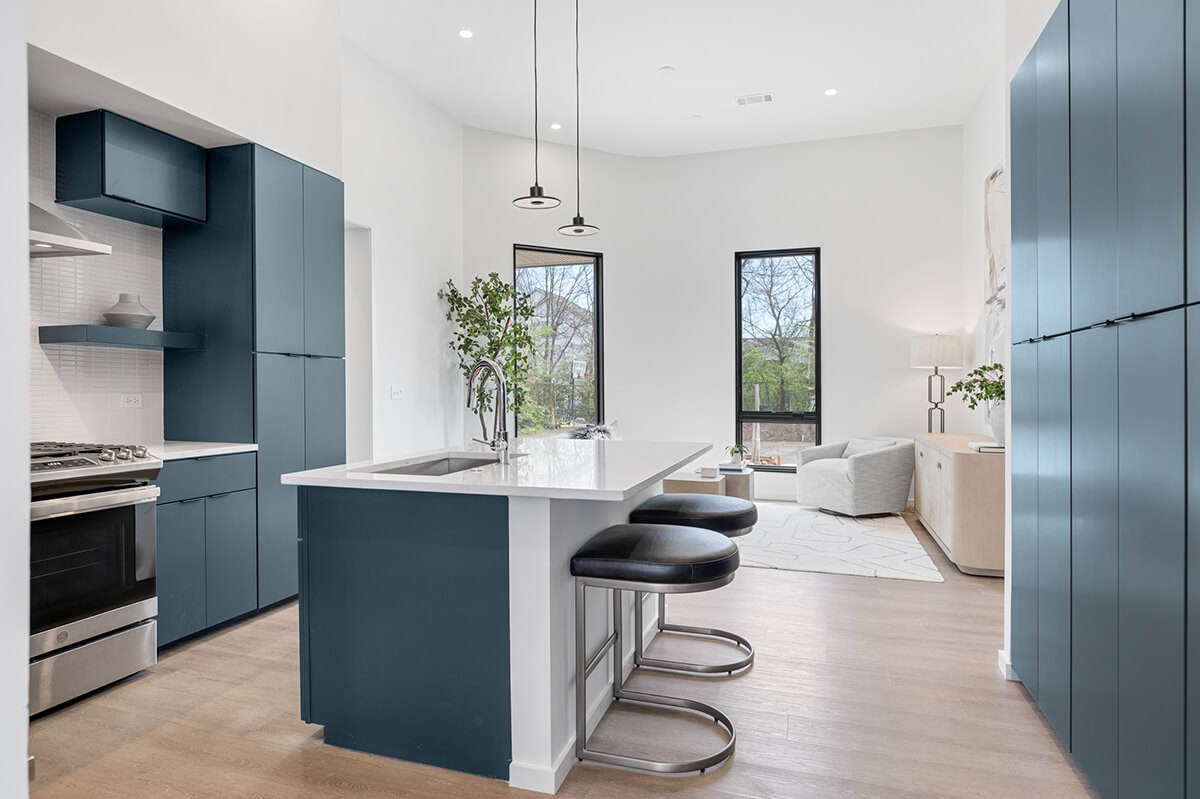 Modern Nashville kitchen at 506 Merritt Ave features blue cabinets, a central island with sink, three bar stools, stainless steel appliances, and an adjacent living area with large windows and neutral decor.