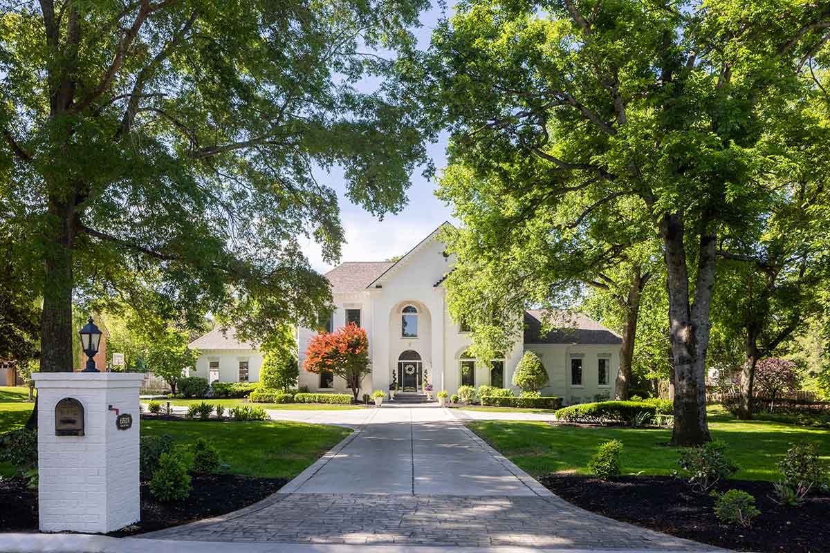 Large white house with arched entryway and manicured lawn, surrounded by tall trees and landscaping, showcases JASPER Builds' exceptional construction, viewed from the end of a driveway on a sunny day.