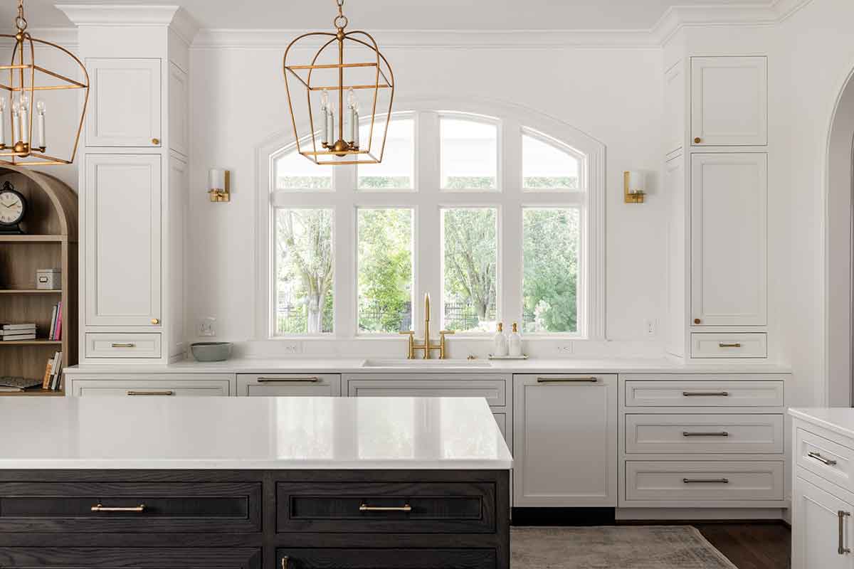 Modern kitchen by JASPER Builds featuring white cabinets, gold hardware, large windows, a white island countertop, and two gold pendant lights hanging from the ceiling.