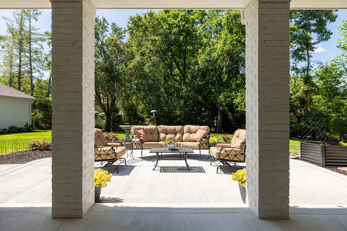 A patio with beige cushioned outdoor furniture and a coffee table sits under a covered area, designed by JASPER Builds, facing a green backyard with trees and gardens.