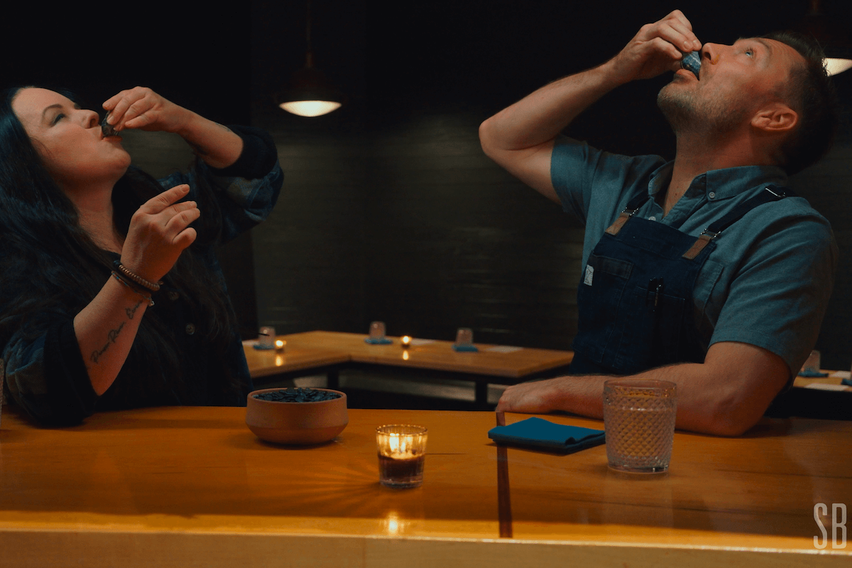 Two people sit at a wooden table in a dimly lit room for StyleBlueprint’s Pull Up a Chair video series, tilting their heads back while eating, with a bowl, a glass of water, and candles set on the table.