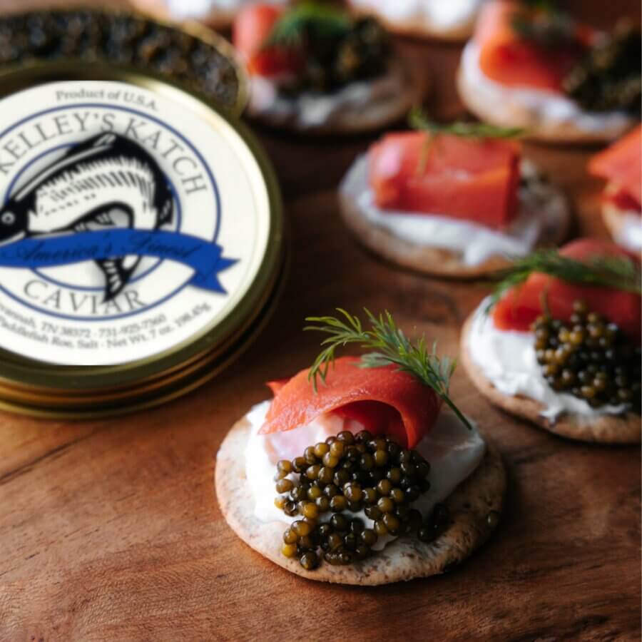 Close-up of crackers topped with cream, smoked salmon, dill, and caviar from one of the finest Southern caviar brands—an open tin of Kelley’s Katch caviar sits on a wooden surface.