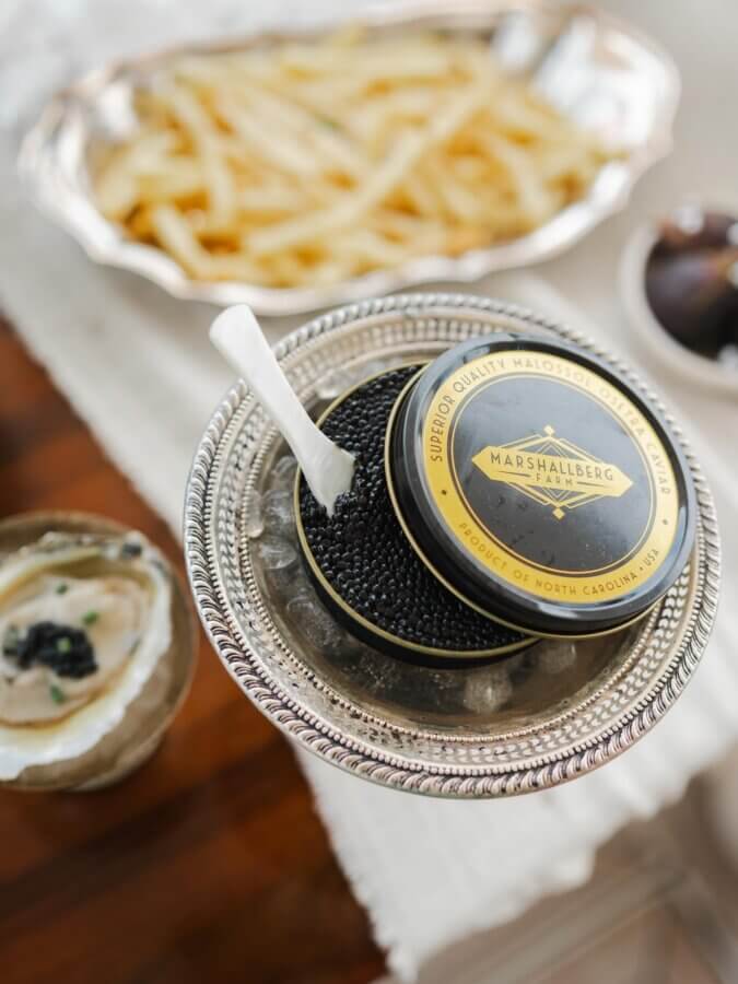A tin of Marshallberg caviar from top Southern Caviar Brands rests on ice, served with a white spoon, crispy fries, and a small dish of creamy spread topped with caviar.