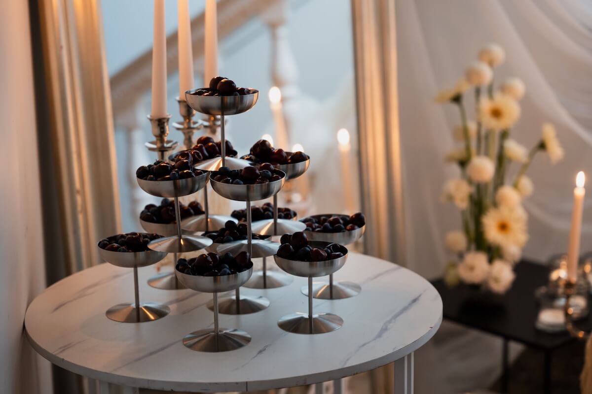 A white round table with tiered metal stands holding cherries, surrounded by lit candles and flowers in a softly lit room, captures one of this year's most romantic wedding trends.
