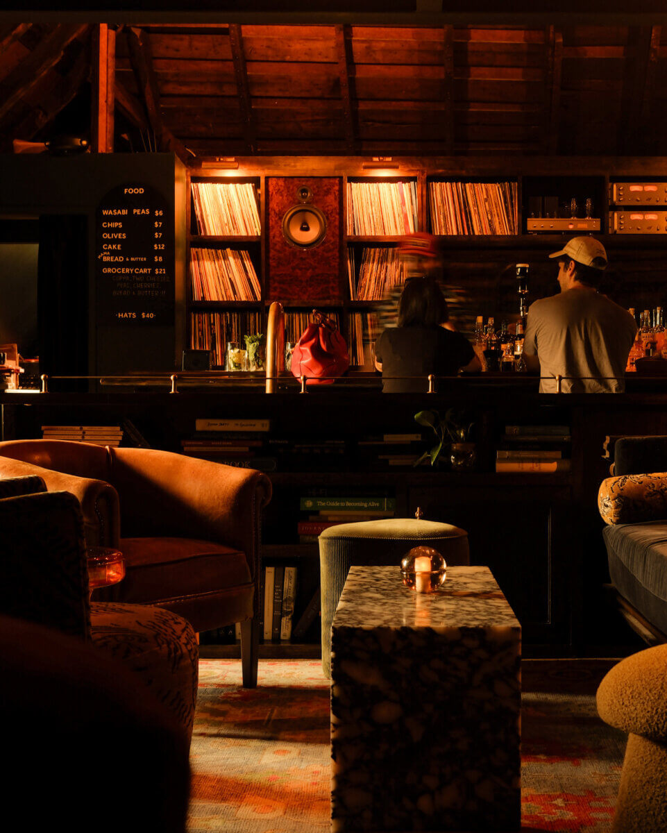 Two people sit at a dimly lit vinyl lounge bar with shelves of books and records, cozy armchairs, a marble table, and a glowing candle in the foreground.