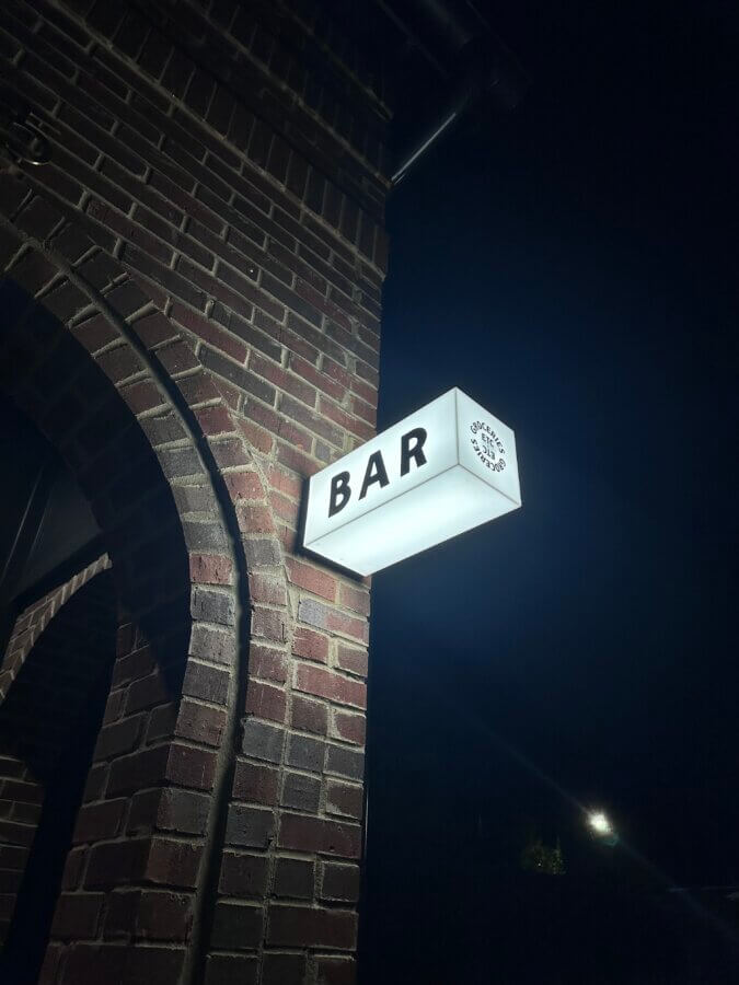 The Rise of Nashville's Vinyl Lounges: 3 to Visit Now + 1 Coming Soon - 3 A lit white sign reading "BAR" is mounted on a brick building at night, hinting at a cozy vinyl lounge inside, with a dark sky and streetlight visible in the background.