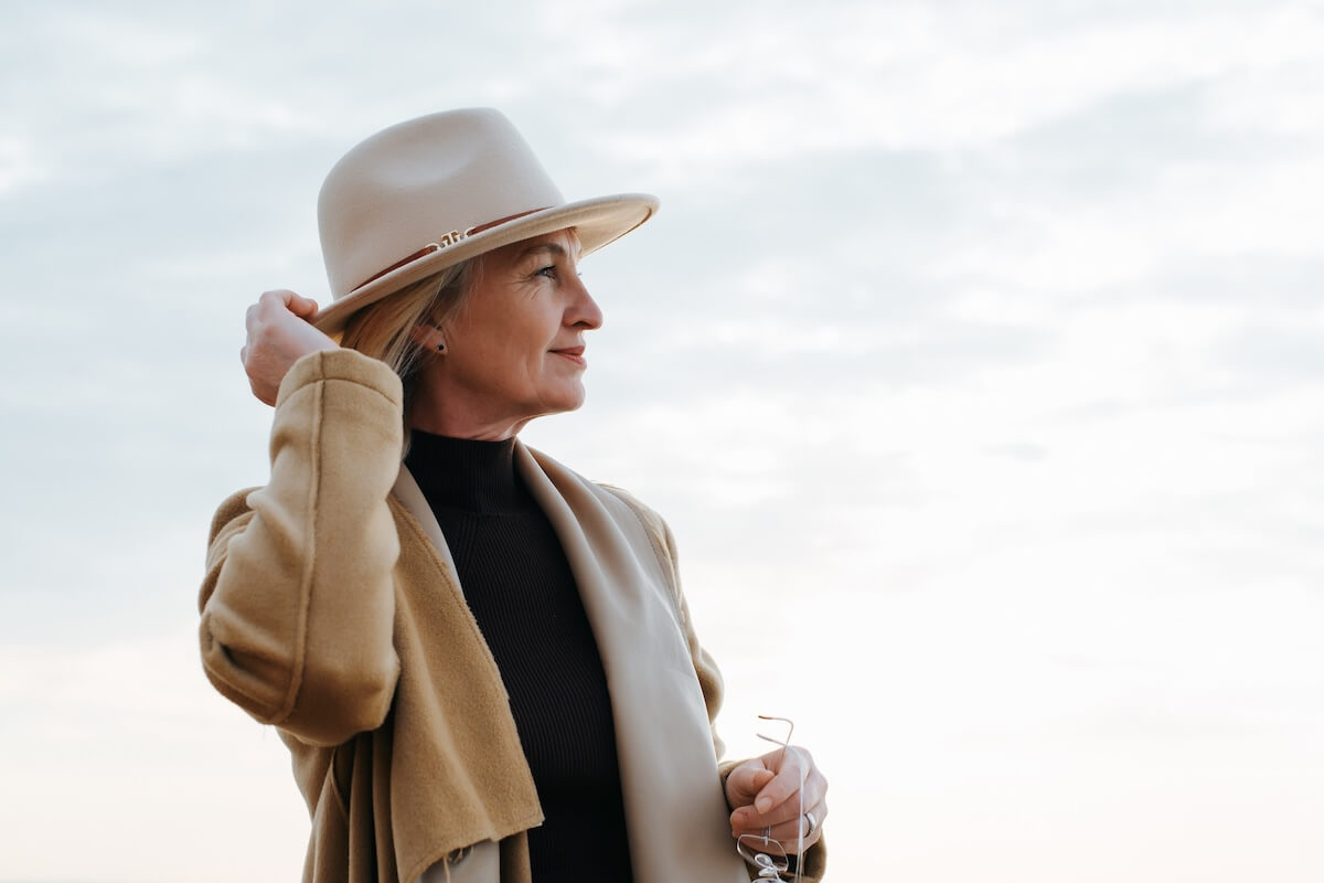A woman wearing a beige coat and crushable hat stands outdoors, looking to the side and holding eyeglasses in one hand against a cloudy sky background.