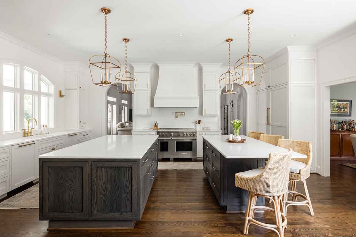 Spacious modern kitchen by JASPER Builds with two large islands, white cabinetry, wicker bar stools, pendant lights, and stainless steel appliances on dark wood flooring.