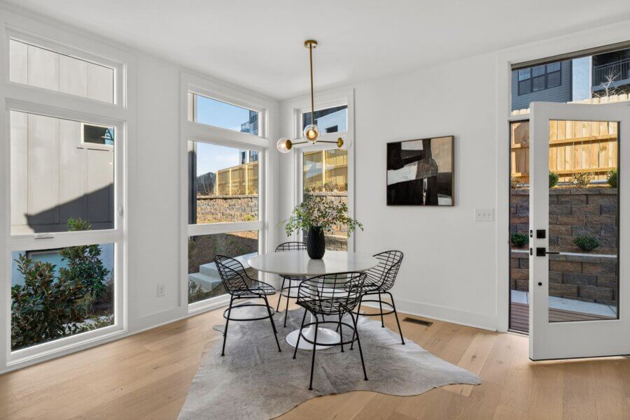 A modern dining nook at 3017 Edwin Circle, Nashville TN, features a round table, four black wire chairs, a cowhide rug, a black vase with greenery, wall art, and large windows overlooking a fenced backyard.