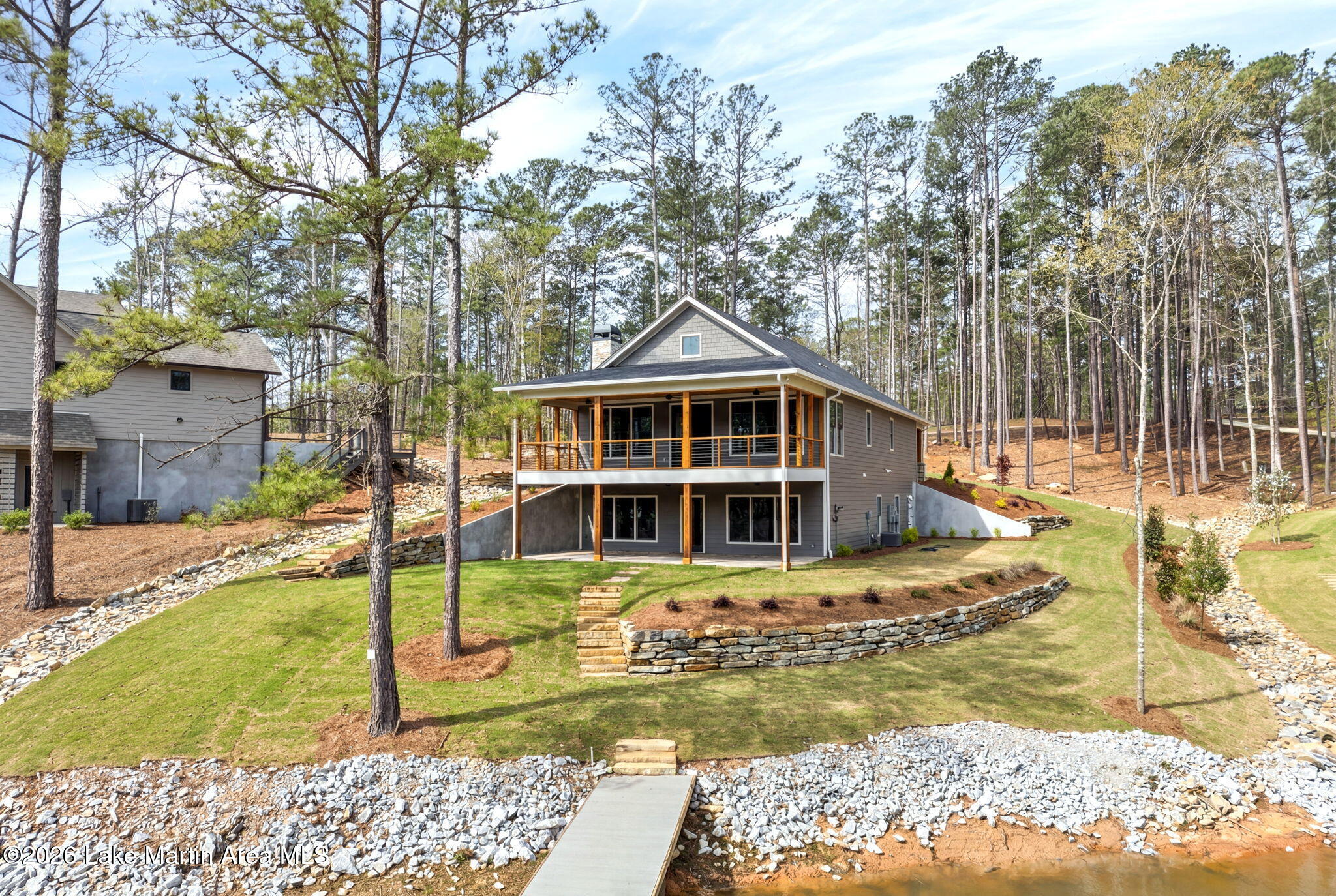 A two-story house at 517 Transom Drive features a large covered porch overlooking a grassy yard and Lake Martin, surrounded by tall trees, beautiful landscaping with stone retaining walls, and a dock leading to the water.