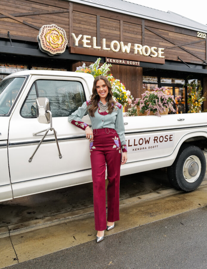 Kendra Scott's Latest Concept Has Landed in Nashville: Apparel, Craft Cocktails & More - 8 A woman stands beside a vintage white truck labeled "Yellow Rose Kendra Scott" in front of the Yellow Rose store in Nashville, decorated with flowers, showcasing a unique retail concept.