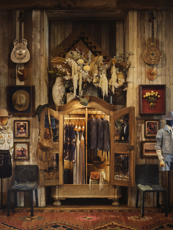 Kendra Scott's Latest Concept Has Landed in Nashville: Apparel, Craft Cocktails & More - 3 A rustic display features a wooden wardrobe with western clothing and accessories, surrounded by guitars, hats, framed art, and dried floral arrangements against a wooden wall.