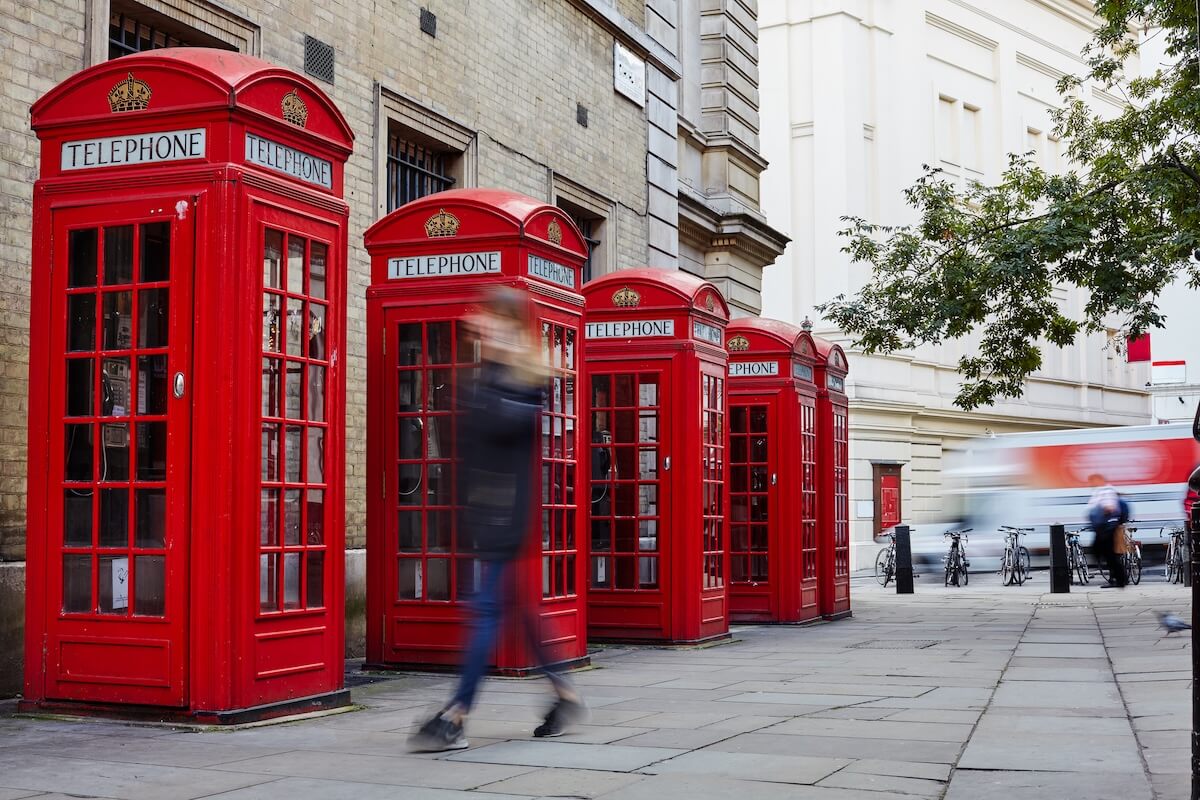 Four classic red British telephone booths line a city sidewalk, adding a touch of Britishisms. A blurred person walks past, with buildings and a tree in the background.