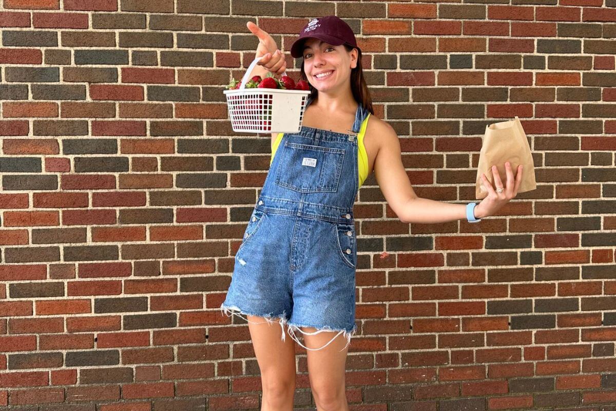 A woman in denim overalls and a maroon cap holds a basket of strawberries and a brown paper bag, standing in front of a brick wall—capturing the charm of One Week in Birmingham.