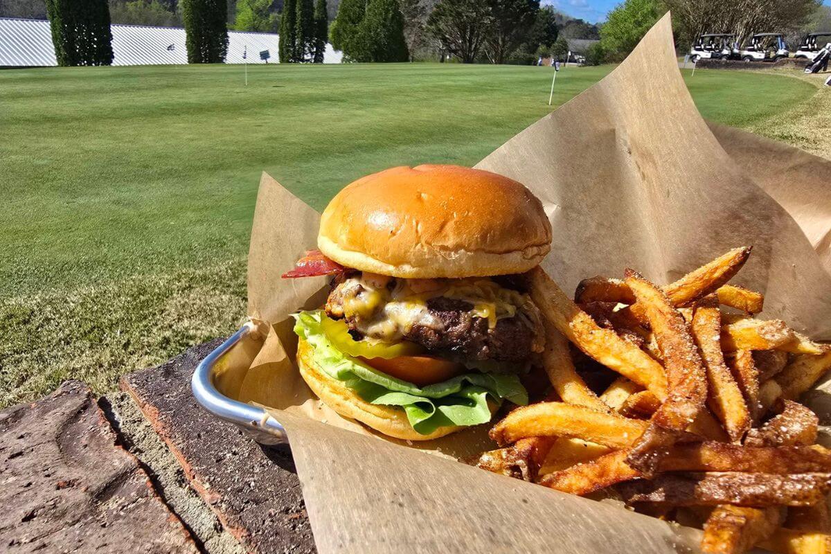 A cheeseburger with lettuce, tomato, and bacon, served with seasoned fries on brown paper near a golf green—a must-try for anyone exploring new restaurants in Birmingham.